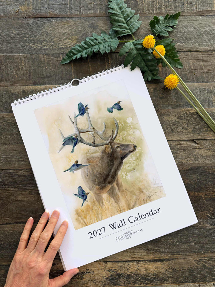 2027 wildlife watercolor calendar cover featuring an elk with cowbirds, displayed on wood background with dried flowers