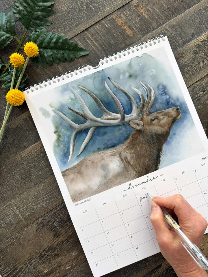Hand writing an appointment on the December page of the 2027 wildlife watercolor calendar, featuring a bull elk painting